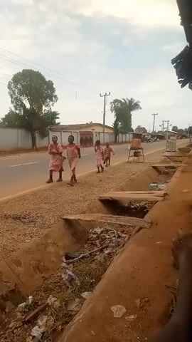 A group of four children, dressed in matching pink and white patterned school uniforms, are walking along a dusty roadside next to a paved road. The children appear to be between the ages of 6 and 10. Two of the children are walking closely together, and one of them is carrying a backpack. Another child is walking a short distance behind them, and the fourth child is further ahead, walking alone. A red and black motorcycle is parked on the side of the road next to the children. The road is bordered by a gravel shoulder, and a drainage ditch filled with debris is visible in the foreground. In the background, there are buildings, trees, and utility poles. The sky is overcast.