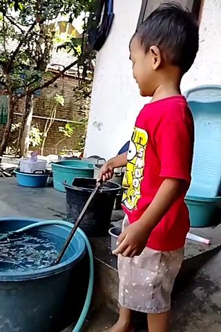 A young boy in a red t-shirt and patterned shorts stands next to a large blue bucket of water. He is holding a wooden stick and appears to be playing with the water. There are several other buckets and containers in the background, as well as some foliage. The scene appears to be outdoors in a backyard or courtyard.