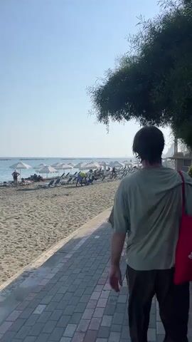 A person is walking away from the camera on a paved path next to a sandy beach. The person is wearing a light green t-shirt, dark pants, and carrying a red bag over their shoulder. The beach has several rows of empty lounge chairs and umbrellas. People can be seen sitting on the chairs and near the water. The sky is clear and blue, and there is a tree with green leaves overhanging the path. The person adjusts their hair as they walk.