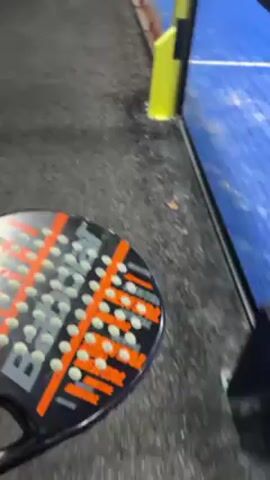 A video shot from behind a chain-link fence reveals a paddle tennis court in an indoor facility. The court is predominantly blue with white lines delineating the playing area. Two people are visible on the court, seemingly engaged in a game or practice. The facility is well-lit with overhead lights. The video appears to be recorded from a stationary position outside the court.