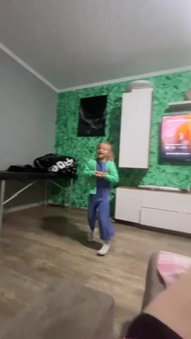 A young girl with blonde hair pulled into a ponytail, wearing a blue jumpsuit and a green zip-up hoodie, dances in a living room. She has white socks on her feet. The room has light wood-paneled floors and gray walls, with one wall covered in a textured green material resembling moss or foliage. To the left, a black massage table is visible with a black bag on it. Above the table, a black rectangular artwork hangs on the gray wall. To the right, a white cabinet is mounted on the green wall, and a television screen is visible, displaying an image. A gray couch is partially visible in the foreground on the right. The girl performs various dance moves, including arm gestures and body movements, facing the camera.