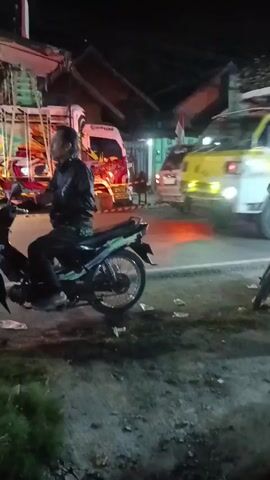 At night, a person is riding a motorcycle on the side of a road. The person is wearing a dark jacket and dark pants. The motorcycle is black with green accents. In the background, there is a truck with a yellow and white cab and a load of what appears to be wood on its trailer. The truck is moving. There are buildings and other vehicles, including more motorcycles, on the side of the road. There is trash on the ground near the motorcycle. The scene is illuminated by streetlights and the headlights of vehicles.