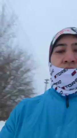 A man wearing a light blue jacket, a patterned beanie, and a white gaiter with the word "TORERO" printed on it, is standing in a snowy outdoor environment. He is facing the camera and appears to be recording a video. Behind him is a snow-covered landscape with trees and an old wooden house with a roof heavily covered in snow. The sky is overcast, suggesting it might be snowing or has recently snowed. The man remains in the same spot throughout the video, moving slightly, and the camera focuses on him and the snowy scene in the background.