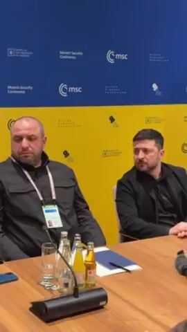 Two men are seated at a wooden table in front of a backdrop that alternates blue and yellow. The man on the left wears a black button-down shirt, a lanyard around his neck, and has a serious expression. To his right is another man, Volodymyr Zelenskyy, who is also dressed in black. Between them on the table are bottles of water and other beverages. Papers, a cell phone, and a microphone are also present. The backdrop is decorated with logos including "Munich Security Conference," "MSC," and "Viktor Pinchuk Foundation."
