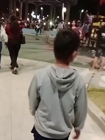 A young boy in a gray hooded sweatshirt and dark pants stands facing away from the camera, looking up towards the right. The boy is in a public outdoor space at night. In the background, there are lights, several other people, and what appears to be a playground structure on the right side of the frame. The ground is paved.