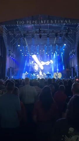 This is a video of a live music performance at the Tio Pepe Festival. The stage is set up with a large screen in the background displaying a live feed of a man playing a guitar. There are several spotlights shining on the stage, illuminating the band members and the screen. A large crowd of people are visible in the foreground, facing the stage and watching the performance. The lighting is predominantly blue, creating a vibrant atmosphere. The overall scene captures the energy and excitement of a live concert.