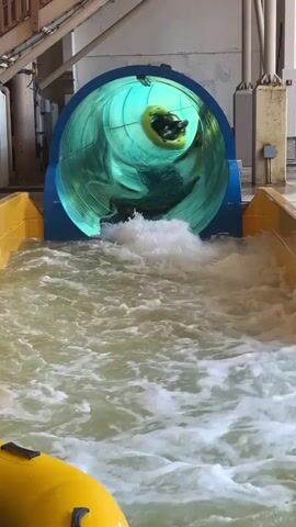Two people are riding a yellow raft down a water slide. The raft is entering a large, blue, translucent tube that is illuminated with a green light. The people are wearing dark clothing. There is a lot of water splashing around the raft and the slide. The environment appears to be an indoor water park.