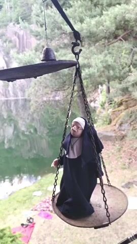 A video shows a close-up, slightly high-angle shot of an antique-looking balance scale set outdoors. The scale has two large, circular metal pans suspended by chains from a central beam. On the right-hand pan, a small figurine of a nun is seated, dressed in a black habit and white wimple, with what appears to be a small white cigarette in its mouth. The nun figurine is facing slightly to the left. On the left-hand pan, a small, dark, bell-shaped object rests. The scale appears to be slightly tipped, with the pan holding the nun figurine lower than the pan with the bell. The background features a calm body of water, possibly a lake or quarry pond, which reflects the surrounding green trees and a rocky cliff face. The lighting is soft and natural.