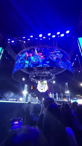 The video shows a lively outdoor concert or festival with a large crowd. The stage is illuminated with bright blue and white lights, and a large circular screen above the stage displays images of people dancing and interacting. There is also a disco ball hanging from the ceiling. The crowd is diverse, with people of various ages and ethnicities. Many are seen holding up their phones to record the event. The overall atmosphere is energetic and celebratory, with people dancing, cheering, and enjoying themselves. Some individuals are seen interacting with each other, taking photos, and smiling. The lighting is dynamic and contributes to the festive mood. The environment appears to be an open-air venue, possibly at night, given the darkness in the background.