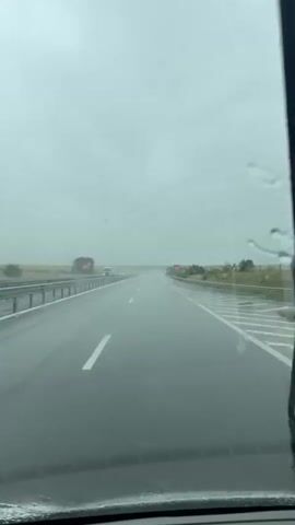 This is a first-person perspective video of driving on a wet highway during a cloudy day. The road is wet with visible reflections of the sky and surrounding environment. The sky is overcast and grey, suggesting rain or heavy cloud cover. Lane markers and a guardrail are visible along the side of the road. There are a few vehicles in the distance, including trucks and cars. The overall atmosphere is subdued due to the weather conditions.