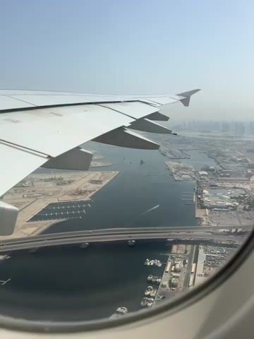 The video shows an aerial view from an airplane window. The wing of the plane is visible in the foreground, taking up the left side of the frame. Below, a body of water is seen with several boats docked and moving within it. A bridge crosses over a section of the water. Beyond the water, there is a sprawling cityscape with numerous buildings, some appearing to be under construction. The sky is hazy.