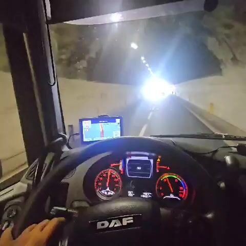The video shows a first-person view from inside a truck cab. The driver's hands are visible on the steering wheel. The truck is driving through a tunnel, with bright lights visible at the end. A GPS device is mounted on the dashboard. As the truck exits the tunnel, the scene transitions to a highway with other vehicles visible. The truck then continues driving on the highway, with a car visible in front of it.