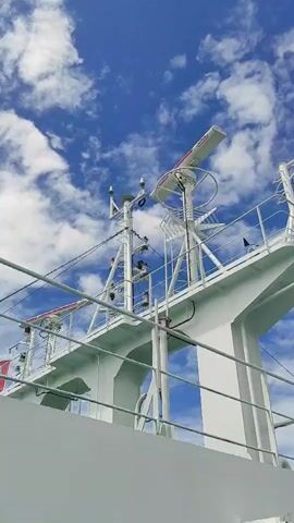The video shows a close-up, low-angle shot of the upper structure of a white ship against a bright blue sky with scattered white clouds. The ship's superstructure is predominantly white and features various antennas and radar equipment, including a prominent rotating radar dish. Metal railings and cables are visible, suggesting a deck area. The camera angle emphasizes the height and complexity of the ship's navigation and communication systems. There are no people visible in the frame. The environment is outdoor, likely at sea, given the context of a ship and the sky.
