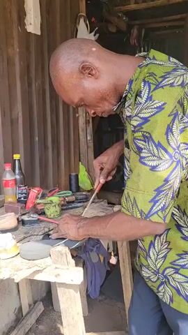 A man in a brightly colored shirt is working on a shoe. He is bald and appears to be concentrating on his task. He is holding a tool, possibly a knife or awl, and is using it to work on the sole of the shoe. The shoe is dark and appears to be a sneaker or athletic shoe. The man is working at a makeshift workbench, which is made of wood and appears to be in an outdoor or semi-outdoor setting. There are various tools and materials on the workbench, including bottles, cans, and other shoe-making supplies. The background shows a corrugated metal wall and some wooden supports, suggesting a rustic or informal workshop. The lighting is natural and appears to be coming from the side.