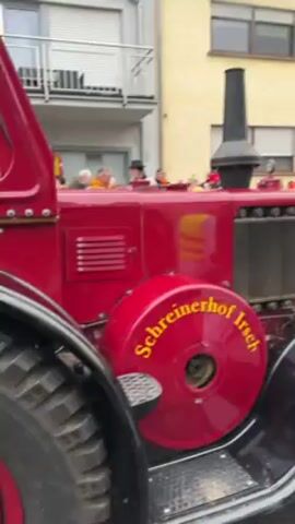 The video shows a vintage red tractor with "Schreinerhof Irsch" written on the wheel. The tractor is pulling a float in a parade, with people in costume standing on the float. The crowd along the street is also dressed in costume. The float is decorated with Asian-themed flags and has writing on the side including "JONAH" and "SUSHI KIMONO IRSCH ALAAF ALAAF". The weather appears to be overcast.