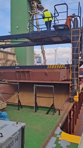 The video depicts a loading operation at a port. Two individuals, one in a yellow safety vest and another in orange, are on an elevated platform overseeing the loading of grain (or a similar granular substance) into a cargo hold of a barge. The loading apparatus consists of a structure extending from a tall, green industrial building. A mechanical arm delivers the grain into the hold below. The hold is mostly full.  A ladder provides access to the platform. Wind turbines are visible in the distance. A crane is also visible in the distance. The deck of the barge is painted green and has various fixtures and equipment on it. The day appears to be somewhat overcast.