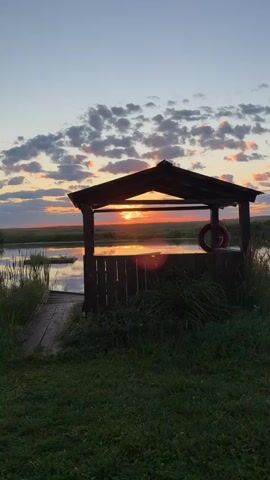 A wooden gazebo with a dark roof and weathered wooden walls stands on the right side of the frame, partially obscuring the setting sun. A red life preserver is attached to the right-hand wall of the gazebo. In front of the gazebo is a calm body of water reflecting the sky, with reeds and tall grass growing on its banks. A wooden plank walkway leads from the grassy foreground into the water, ending near the gazebo. The sky is a gradient of blue and orange, with fluffy clouds catching the last rays of the sun. The overall scene depicts a peaceful sunset over a natural landscape.