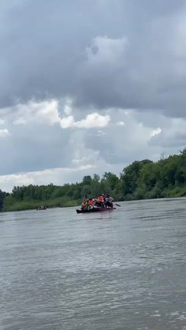 The video shows a wide, murky river under a cloudy sky. On the river, a raft is carrying several people, most of whom appear to be wearing orange life vests. The raft is moving away from the camera, towards the left side of the frame. In the background, a dense line of green trees runs along the riverbank. The sky is filled with heavy, dark gray clouds, suggesting an overcast or stormy day. There is another, smaller raft visible in the distance to the left of the main raft. The water appears to be moving, with ripples and waves on the surface.