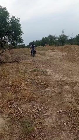 A person rides a dirt bike on a dry, dusty, and overgrown path. The rider, wearing a helmet and dark clothing, is positioned in the center of the frame. The path is surrounded by trees and dry vegetation. The sky is overcast.
