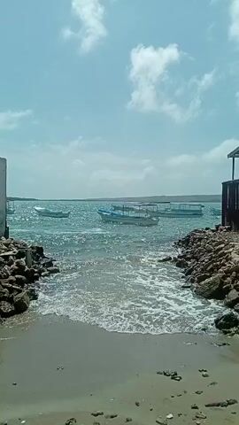 A video shows a body of water on a sunny day. The camera appears to be at the edge of a small beach, between two small rock formations, looking out over the water. Several small boats are visible on the water, mostly in the center of the frame, and appear to be gently moving with the waves. The water is a bright blue-green color. The sky is blue with some white clouds. There is a small building on the right side of the frame. The water is rough and choppy, and small waves are breaking on the shore. There is a voice in the background.