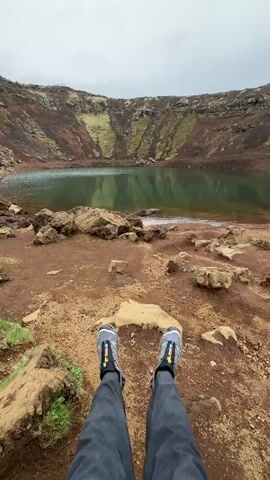 The video shows a first-person perspective of someone sitting on the edge of a volcanic crater, with a lake at the bottom. The person is wearing gray pants and gray and black hiking shoes. The ground is reddish-brown and rocky with patches of green vegetation. The crater walls are multicolored with layers of red, brown, and green. The lake water is green and reflects the sky. The sky is overcast. In the distance, figures of people can be seen walking along the crater rim.
