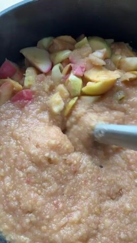 The video shows a close-up shot of an apple puree being blended in a pot. There are pieces of apple on top of the puree. A blender is being used to blend the apples. The pot is black and appears to be made of metal. The lighting is bright, suggesting the video was taken during the day. The overall scene is one of food preparation.