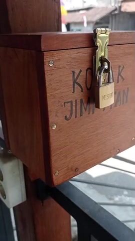 The video is a close-up, static shot of a wooden box labeled "KOTAK JIMATAN" (Savings Box) with a small padlock securing it. The box is attached to a wooden post. In the background, out of focus, is a street scene with buildings and a vehicle. There are no people or actions depicted in the video.