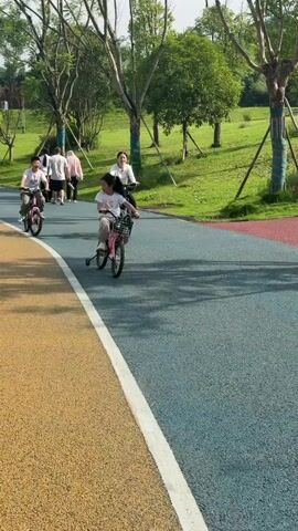A group of people are riding bicycles on a paved path in a park. The path is multi-colored, with sections of yellow, blue, and red. There are trees and green grass on either side of the path. The people are mostly children, dressed in casual clothing. They are all looking forward and appear to be enjoying themselves. One of the children is in the foreground, riding a pink bicycle with training wheels. Another child is behind her, also on a pink bicycle. Further back, an adult is riding a black bicycle. The sun is shining, casting shadows on the path.