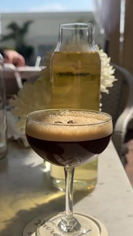 The video shows a sunny outdoor patio with a marble-patterned table. In focus is an espresso martini in a stemmed glass, garnished with three coffee beans on top of its foamy head. To its left, there's an empty glass carafe, and to its right, a taller carafe filled with a light yellow liquid, possibly lemonade or iced tea, next to a white flower arrangement. In the background, blurred figures are visible, including a person sunbathing on a lounge chair. The sky is blue and clear. The scene appears to be a relaxed, upscale outdoor setting.