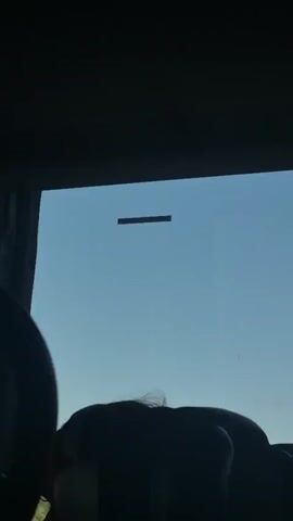 The video shows a view from inside a vehicle looking out a window. The foreground is dark, obscuring details of the vehicle's interior. The background shows a bright blue sky with a dark horizontal object floating in the distance. Trees and foliage are visible at the very beginning of the video, suggesting the vehicle is moving through a wooded area. The horizontal object appears to be stationary against the moving background of trees and sky.