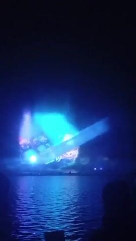 This is a video of a nighttime water and light show. The show features projections of animated vehicles, including cars, trucks, and what appears to be a bus, onto a screen of mist or water. Colorful lights, primarily green, red, and purple, are projected from behind the mist, creating a dynamic and vibrant display. Fountains of water are also integrated into the show, rising and falling in sync with the lights and projections. The scene takes place over a body of water, likely a lake or river, and reflections of the lights are visible on the water's surface. In the foreground, the dark silhouettes of several people are visible, suggesting they are audience members watching the performance. The overall atmosphere is one of entertainment and visual spectacle.