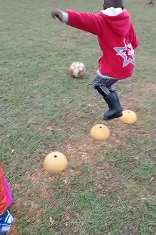 The scene unfolds on a grassy field where two young children are present. One child, dressed in a red jacket with a star design, gray pants, and black boots, is stepping over three yellow half-sphere cones. The second child, in a red jacket and a pink skirt, stands and watches the first child, with what seems to be a bag in hand. A soccer ball lies on the grass between the two children. Other people are visible in the background but are further away. The first child briefly squats and touches one of the cones. The overall setting suggests a casual outdoor activity, possibly practice or play.