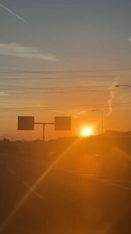 The video shows a car driving on a highway towards a city at sunset. The sun is low on the horizon, casting a warm orange glow over the scene. The highway is mostly empty, with only a few other cars visible. There are streetlights lining the highway. In the distance, the city skyline is visible. The video is shot from the perspective of someone inside the car.
