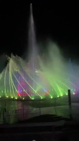 A nighttime view of a musical water fountain show. Water jets are illuminated by colored lights, transitioning through green, blue, pink, and red. The water shoots high into the air, creating a dramatic visual display against the dark sky. The fountain is situated in a body of water, with some lights visible beneath the surface.