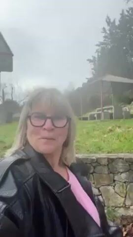 A middle-aged woman with blonde hair and glasses stands outside in front of a stone wall and a grassy area with trees. She is wearing a black leather jacket and a pink shirt. The weather appears to be overcast and misty. The camera seems to be held by the woman. She looks at the camera and smiles. A structure with a roof is visible in the background. There are brown leaves on the ground near the wall. There is a plastic bag by the wall. She pans the camera slightly to the right, showing more of the trees and stone wall in the background.