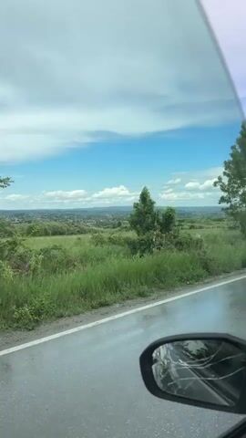 The video, filmed from within a moving vehicle, offers a view of a rural landscape on a day marked by a blue sky interspersed with white clouds. The road is wet, suggesting recent rainfall. Patches of green vegetation and occasional trees flank the roadside. In the distance, a settlement or town can be seen nestled within the landscape. The side mirror of the vehicle is visible, showing cracks on its surface.
