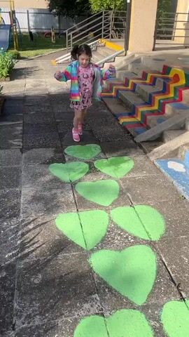 A young girl with dark hair in pigtails, wearing a pink dress with a cartoon print and a colorful striped jacket, is playing hopscotch on a concrete path painted with green heart shapes. She is in mid-jump, with one foot raised. To the right of the path are stairs decorated with colorful rainbow stripes. The girl is facing the stairs and appears to be moving towards them. The environment is an outdoor space, possibly a playground or schoolyard, with some greenery and a building in the background. A shadow of a person holding a phone is visible on the ground to the right of the girl.