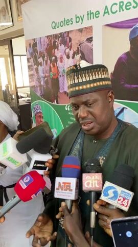 A man in a dark green robe and an ornate hat speaks into a cluster of microphones. The microphones have logos for various Nigerian media outlets. The man is surrounded by people holding the microphones. Behind him is a banner that reads “Quotes by the ACRES.” The banner has images of people, one of whom is in a similar hat. The setting appears to be indoors, possibly a press conference or interview. The man seems to be giving a statement or answering questions from the media. The lighting is bright, and the scene is focused on the man speaking.