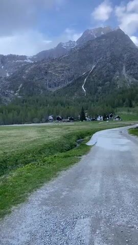 The video shows a mountainous landscape on a cloudy day. In the foreground, there is a gravel path alongside a small stream flowing through a grassy field.  Across the field, there's a line of trees leading to a small collection of buildings with dark roofs and light walls.  In the background, towering mountains are visible, some with patches of snow.  There is a white ski lift infrastructure. The camera pans slightly, revealing more of the valley and mountains.