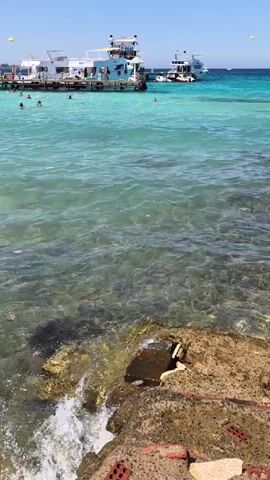 The video shows a sunny day at a tropical beach with clear turquoise water and boats docked in the distance. People are swimming and enjoying the water near the shore. The foreground features a rocky coastline with some debris. The camera appears to be handheld and captures a slight wobble as it pans across the scene.