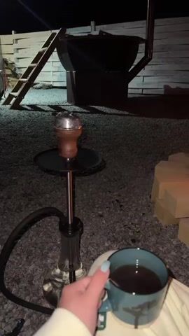 At night, the camera shows a person’s perspective of an outdoor evening scene, seated, with a cup of tea in hand. The foreground features small gravel. To the left, a hookah sits on a round, black surface. A tea mug, teal in color and decorated with a tree motif, is held by a hand with light blue nail polish.  The person wears white comfortable clothing and white slides are visible.  To the right, a circular campfire pit made of beige blocks is lit with a bright, crackling fire. In the background, a large, black cauldron-like structure stands near a white wooden fence with a ladder propped against it.