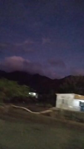 The video shows a blurry, handheld shot of a landscape at dusk or night. The sky is a deep blue with some purple hues and wispy clouds. In the middle ground, there are dark, silhouetted mountains. In the foreground, there are dark trees and some faint lights. A small, white structure is visible on the right side of the frame. The camera appears to be moving, possibly from a vehicle, indicated by the motion blur and shifting perspective. There are no people clearly visible in the footage.