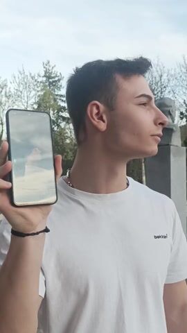 A young man stands outdoors on a partly cloudy day. He wears a white t-shirt, a silver chain, and a black wristband. In his left hand, he holds a black smartphone, which plays a video of himself wearing sunglasses and a white t-shirt. In the background, trees and a monument are visible. A loud, high-pitched noise is audible.