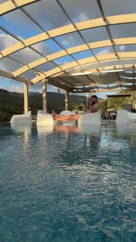 A man sits in a white lounge chair in a swimming pool. He is holding a phone in his left hand and looking at it. The chair is submerged in the water. The pool is covered by a transparent roof with metal beams. In the background, there are more white lounge chairs and lush green hills under a cloudy sky. The water is clear blue with a mosaic tile pattern on the bottom.