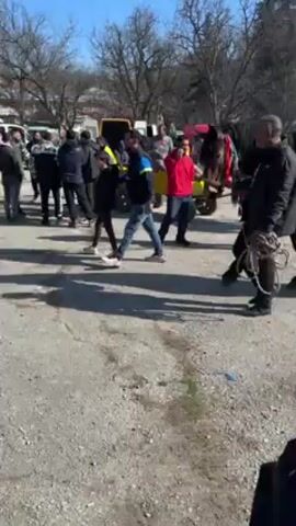 The video shows a group of people standing and walking around in an outdoor area. A horse is harnessed to a yellow cart. The environment appears to be a parking lot or open space with bare trees in the background. There are several vehicles parked in the distance, including vans. The people are dressed in casual clothing. Some are standing and talking, while others are walking. A man holds a coiled rope. The lighting suggests it is daytime.