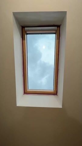 The camera zooms in on a rectangular skylight window surrounded by white walls. The window has a natural wood frame and a partially visible roller shade at the top. Outside the window, the sky is overcast with light grey clouds.