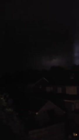The video shows a dark outdoor scene with houses and trees visible. The sky is black, suggesting nighttime. There are no people or animals visible in the frame. The overall lighting is very low, with some faint illumination that may be from distant streetlights or ambient light pollution. The houses appear to be residential structures, and the trees are leafy. There is no discernible action or interaction taking place.