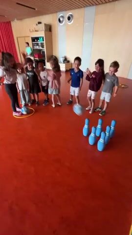 In an indoor setting with vibrant red-orange flooring and light wooden walls, a group of young children, accompanied by an adult, are engaged in a game of bowling. Seven children are prominently featured, standing in a semi-circle around a yellow hula hoop, their eyes fixed on the bowling pins. An adult woman stands to the left, observing and guiding them. In the foreground, several soft blue bowling pins are initially set up, with a small blue ball positioned nearby.

As the video progresses, the ball is rolled from an unseen point, striking and knocking down all the blue pins. Upon witnessing the "strike," the children erupt in joyous cheers and exclamations, raising their arms, jumping excitedly, and clapping their hands. Their faces are alight with happiness and enthusiasm. One child wears a t-shirt with "PARIS" written on it. The scene captures a moment of shared excitement and fun during what appears to be a play activity in a school or daycare environment in La Fouillouse, France, during the daytime.