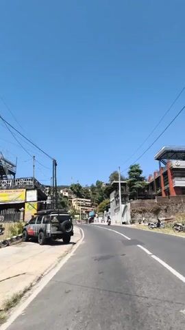 The video shows a road curving uphill with buildings on either side. On the left, there is a parked vehicle, possibly an SUV, with a roof rack and a spare tire mounted on the back. Several motorcycles are parked along the curb to the left of the vehicle. Further up the road, a person is riding a motorcycle. The sky is clear and blue, indicating a sunny day. The buildings appear to be a mix of residential and commercial, with some set against a hillside covered in greenery. Power lines are visible running overhead.
