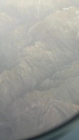 The video shows an aerial view of a mountainous landscape. The mountains are rugged and appear to be covered in dry earth and rocks, with some patches of snow visible on the peaks. The terrain is characterized by steep slopes, deep valleys, and winding riverbeds. The sky above is hazy, with some clouds scattered across it. The overall impression is of a vast and remote wilderness.